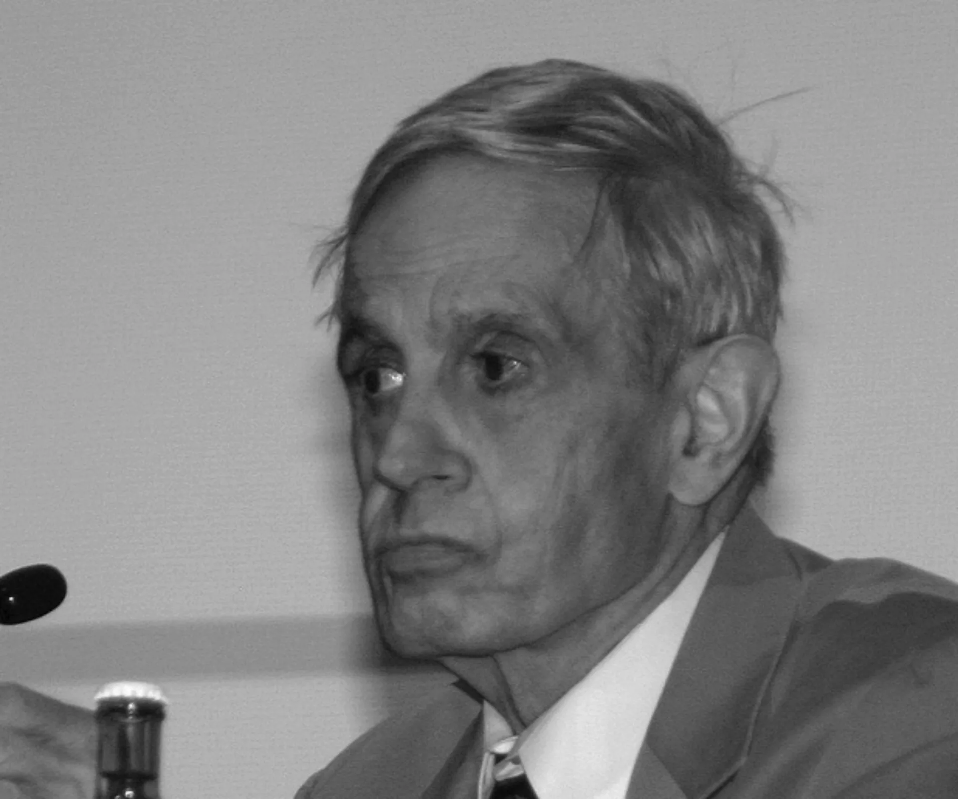 Elderly man with short hair and focused expression, wearing a suit, speaking at a conference with a microphone visible.