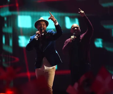 Two performers on stage at Eurovision 2015, one singing with a hat, both holding microphones, vibrant red lighting in background.