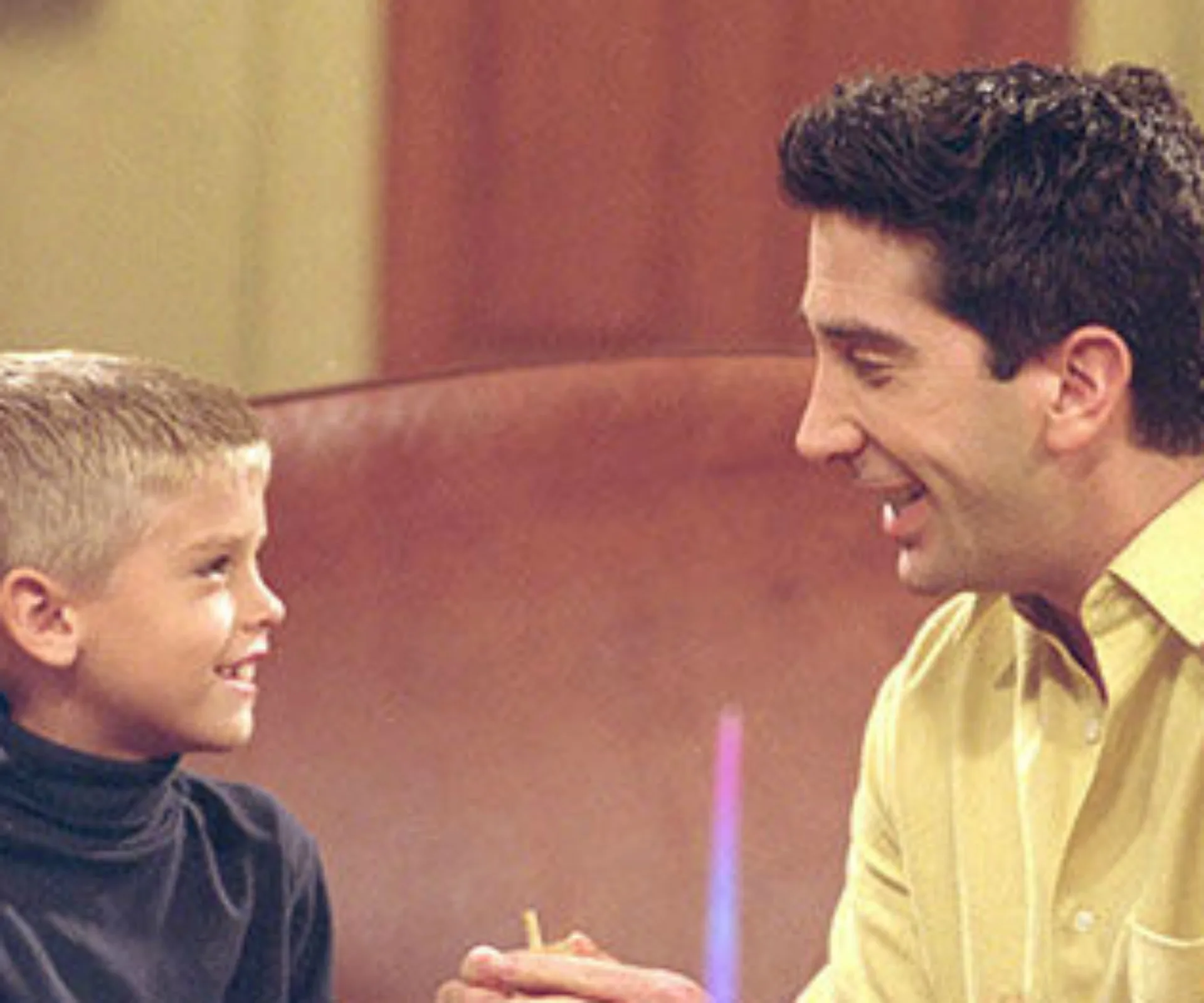 Adult and child smiling at each other while sitting on a couch, in a scene from a TV show.