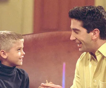 Adult and child smiling at each other while sitting on a couch, in a scene from a TV show.