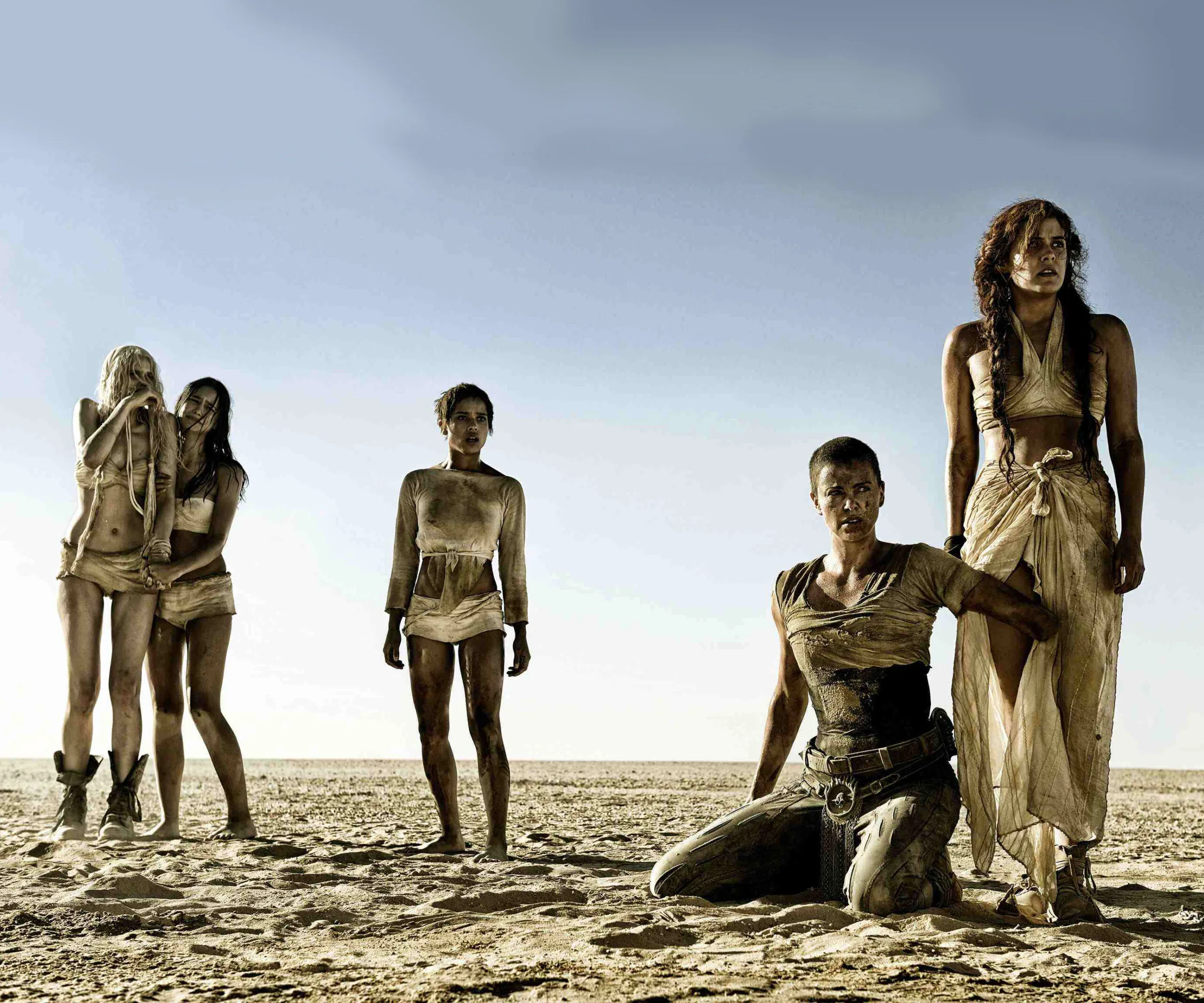 Characters from "Mad Max: Fury Road" stand in a desert, with one kneeling, amid a barren landscape.