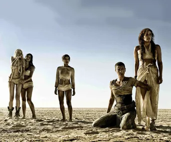 Characters from "Mad Max: Fury Road" stand in a desert, with one kneeling, amid a barren landscape.