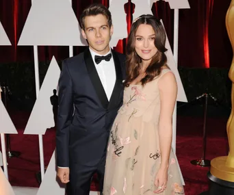A man in a tuxedo poses with a woman in a floral dress on a red carpet event with decorative background elements.