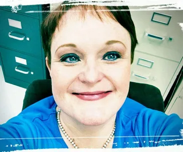 Smiling woman in a blue shirt with a pearl necklace, sitting near filing cabinets.