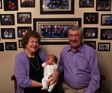 Grandparents smiling with their 100th grandchild, surrounded by framed family photos on the wall.