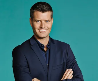 Man in a blue suit smiling with arms crossed, standing against a teal background.