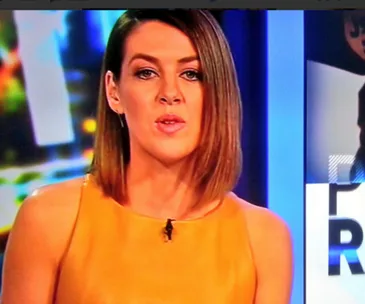 Person with shoulder-length hair in yellow top, speaking on a TV show set.