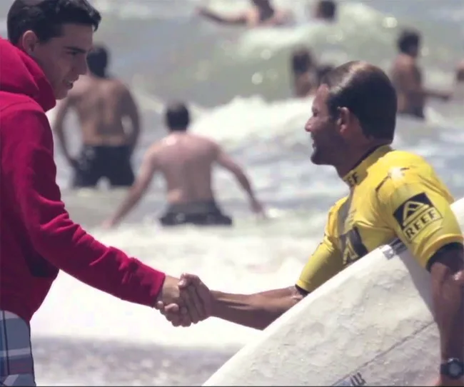 Surfer in yellow rash guard shakes hands with a person in a red hoodie on the beach, surrounded by ocean waves and swimmers.