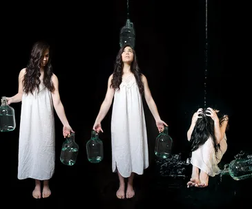 Three women in white dresses holding water jugs, with water pouring over the third woman crouched under the stream.