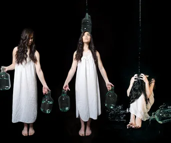 Three women in white dresses holding water jugs, with water pouring over the third woman crouched under the stream.