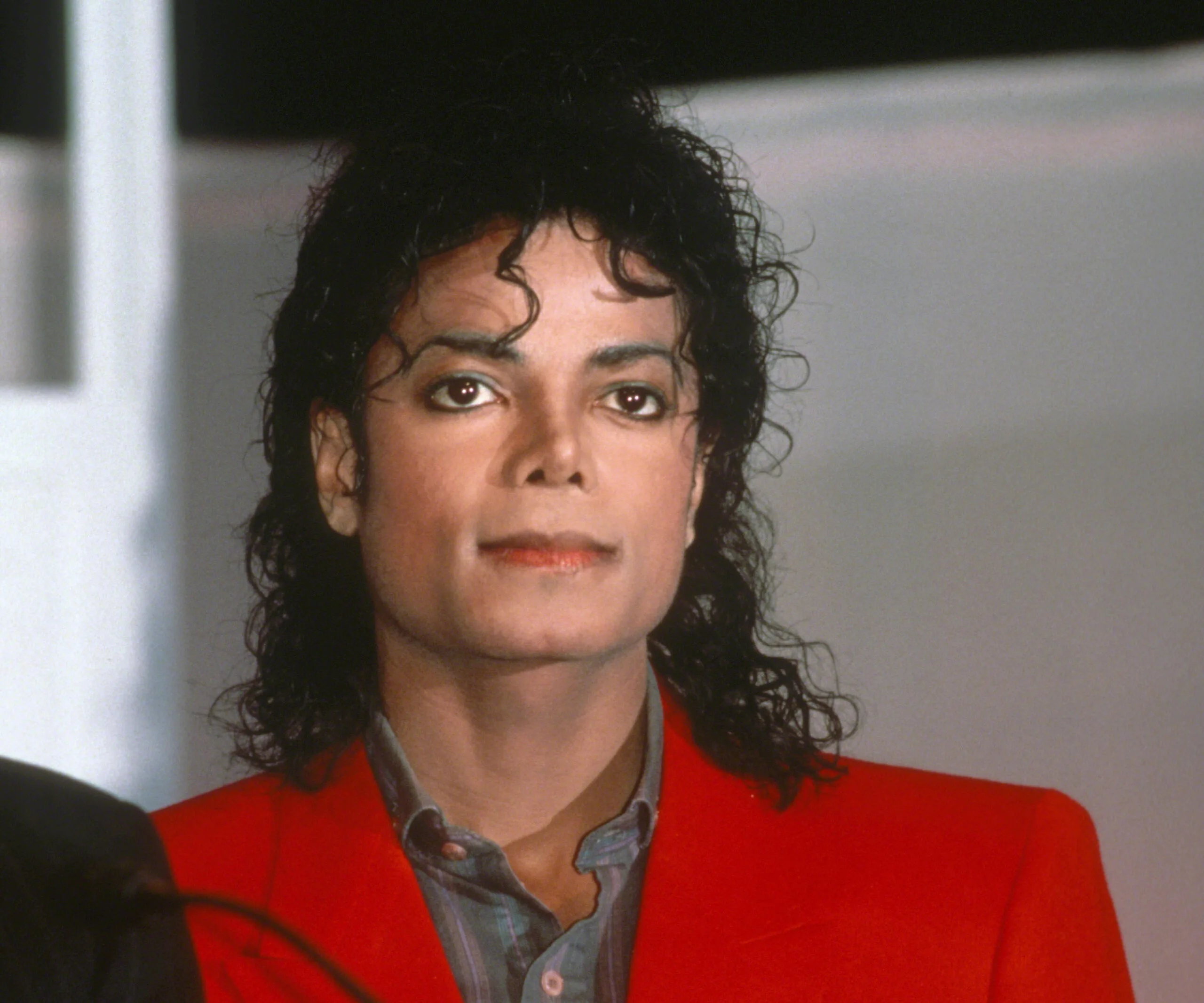 A person with dark curly hair in a red jacket, appearing serious, at an event.