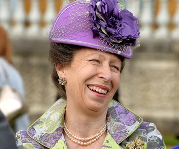 Princess Anne smiling at a royal garden party, wearing a purple hat and floral outfit.