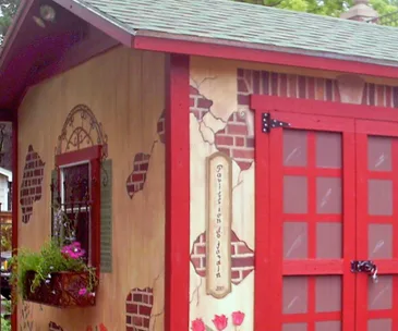 Decorative she-shed with red trim, faux brick design, flower box, and a sign reading "Pavillon de Jardin."