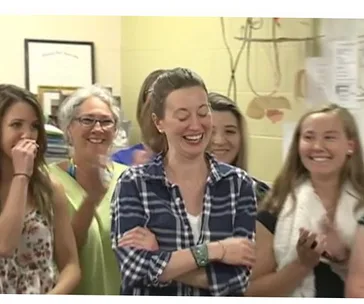 Group of smiling women in a classroom, one in a plaid shirt is in focus, appearing joyful amidst clapping and laughter.