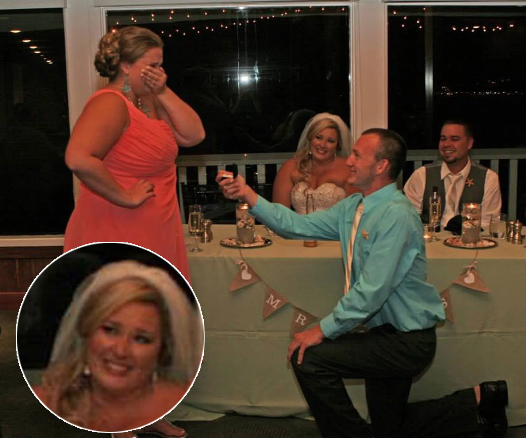 A man proposes to a surprised woman at a wedding reception as two seated guests look on, smiling.