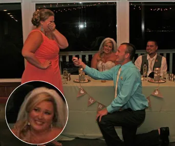 A man proposes to a surprised woman at a wedding reception as two seated guests look on, smiling.