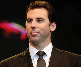 A man in a suit and tie stands on stage with a focused expression, colorful lights in the background.