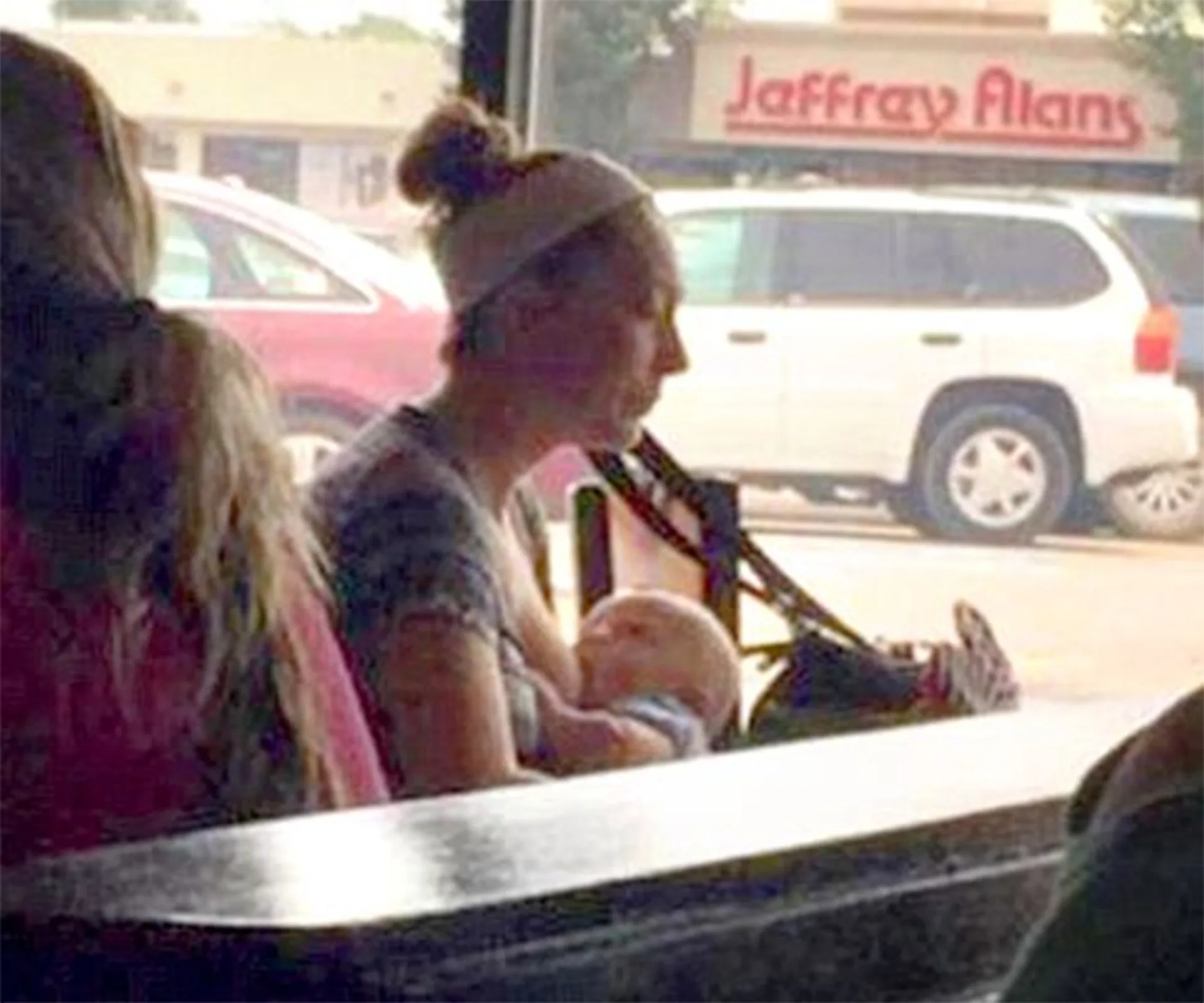 A woman breastfeeding her baby at a cafe with a shopping center visible outside the window.