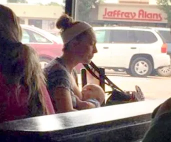 A woman breastfeeding her baby at a cafe with a shopping center visible outside the window.