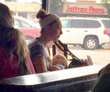 A woman breastfeeding her baby at a cafe with a shopping center visible outside the window.