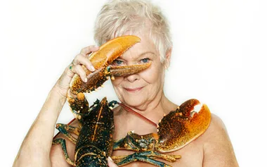 Elderly woman humorously posing with a large lobster, using one claw to playfully cover one eye.