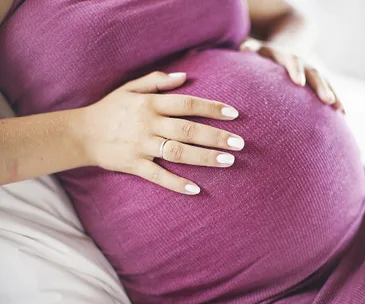 A pregnant woman in a pink dress gently cradles her belly with both hands, showing a white manicure.