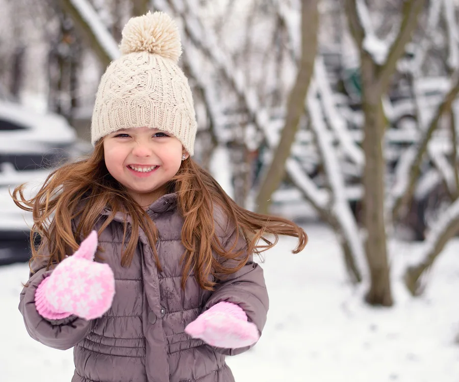 Smiling child in winter coat and hat gives thumbs up in snowy outdoors.