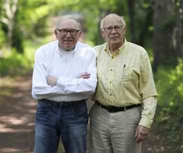 Two elderly men smiling and standing side by side on a wooded path, dressed casually in shirts and glasses.
