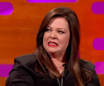Melissa McCarthy looking surprised on a talk show set with a vibrant orange and purple background.