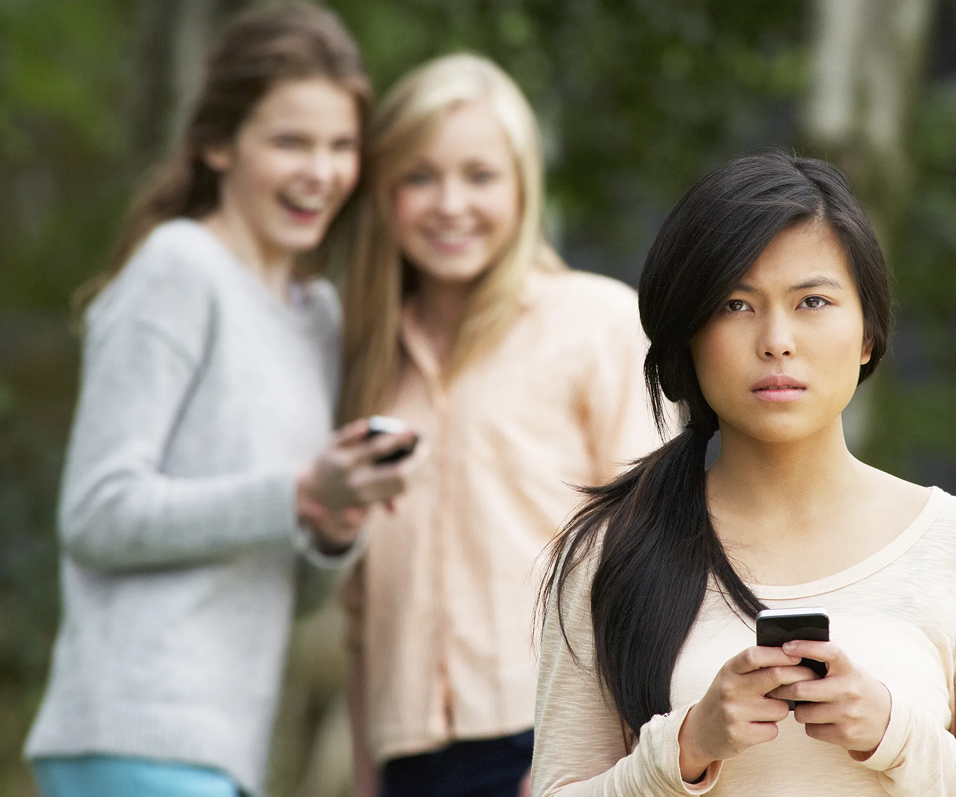 A serious girl with a phone in the foreground; two laughing girls with phones in the background, outdoors.