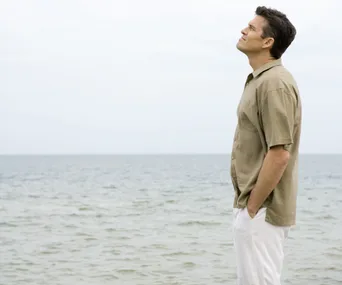 Man in a beige shirt gazing thoughtfully at the sea, hands in pockets, against a gray sky.