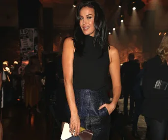 Woman in a black sleeveless top and shiny pants, smiling at an event with dim lighting and people in the background.