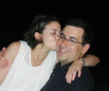 A woman kisses a smiling man on the cheek against a dark background.