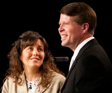 A man in a suit smiles while standing beside a woman with curly hair looking at him, both against a dark background.