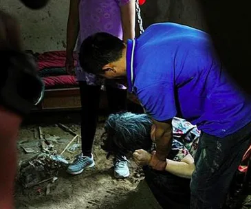 A person helps an elderly woman, who is crouched and chained, inside a dimly lit room with a bed in the background.