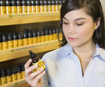 A woman examines a bottle of homeopathic remedy in a store with shelves of similar bottles in the background.