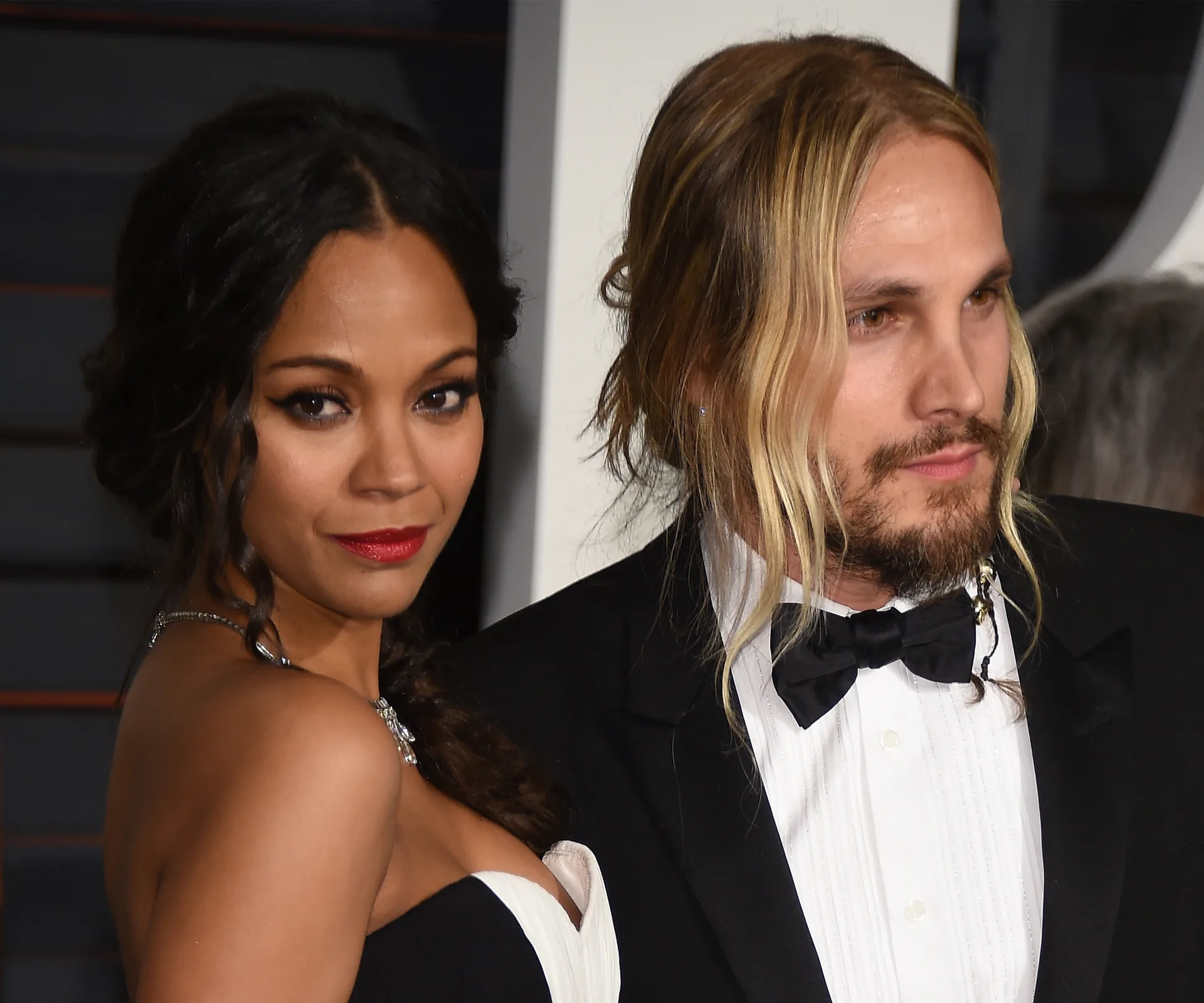 Zoe Saldana in a black and white gown with her husband at a formal event, both looking towards the camera.