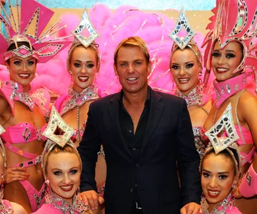Man in black suit surrounded by six women in vibrant pink show costumes with elaborate headpieces.