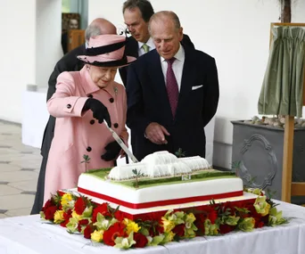 A woman in a pink coat cuts a large decorated cake, surrounded by men in suits, on a table with flowers.