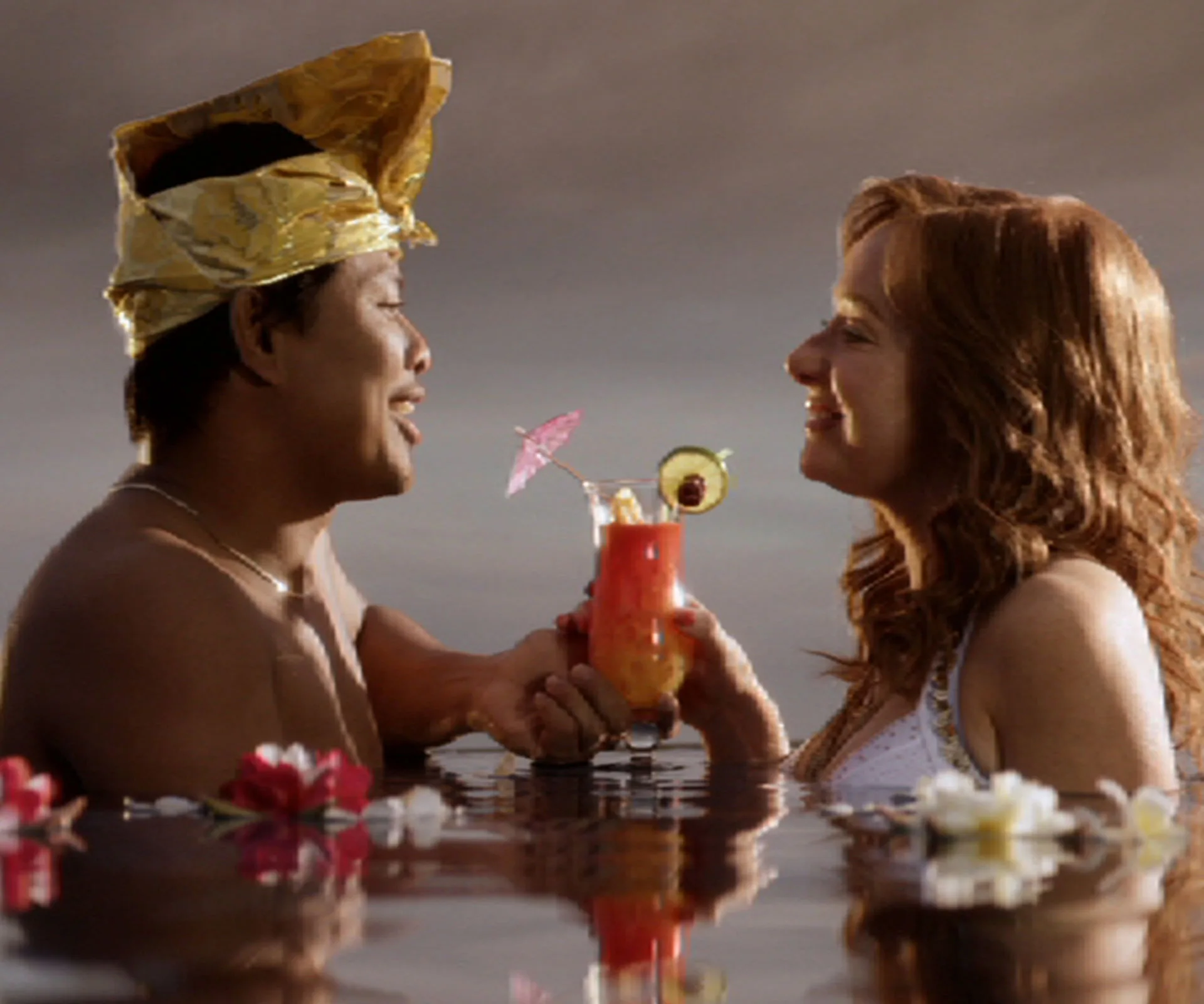 A couple in water, exchanging a drink with a cocktail umbrella, surrounded by floating flowers.