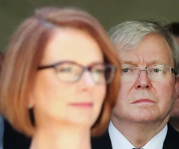 Two individuals in focus, the woman at the front blurred; both wearing glasses, expression serious, outdoor setting.