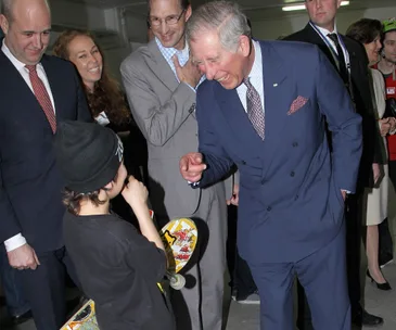 A man in a suit laughing and interacting with a child holding a skateboard during a lively group gathering.