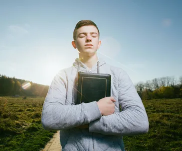 A person outdoors with eyes closed holds a book against their chest, standing in a field with sunlight in the background.