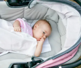 A baby in a pink onesie lies in a stroller, covered with a white blanket, looking to the side.