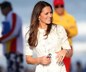 Woman in a white dress smiling outdoors, holding sunglasses.