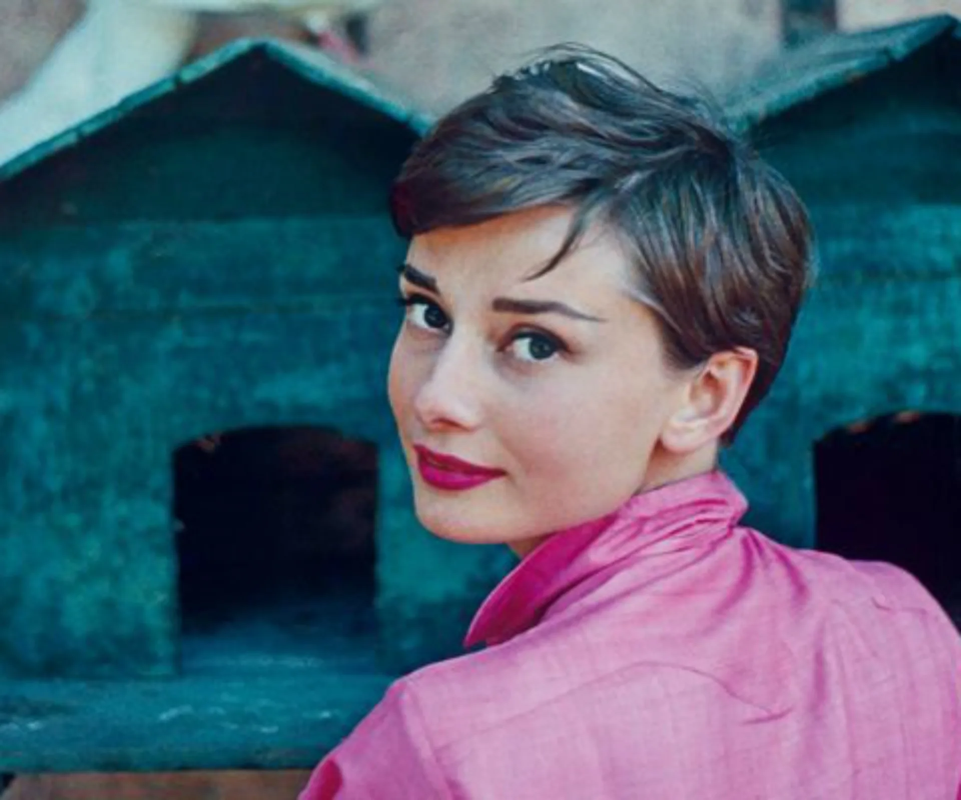 Classic portrait of Audrey Hepburn in a pink shirt with short hair, turning to face the camera, vintage setting.