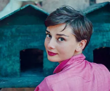 Classic portrait of Audrey Hepburn in a pink shirt with short hair, turning to face the camera, vintage setting.