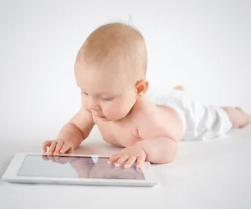 A baby lying on their stomach, interacting with a tablet on a white background.