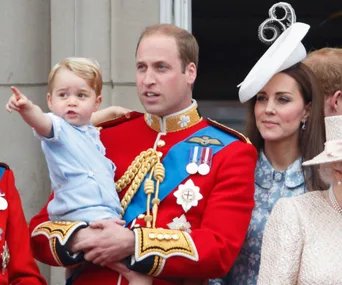 A child pointing, held by a man in red, with a woman beside them wearing a hat, possibly at an official event.