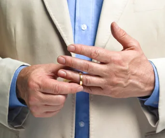 Hands of a person in a beige blazer, removing a wedding ring, with a blue shirt underneath.